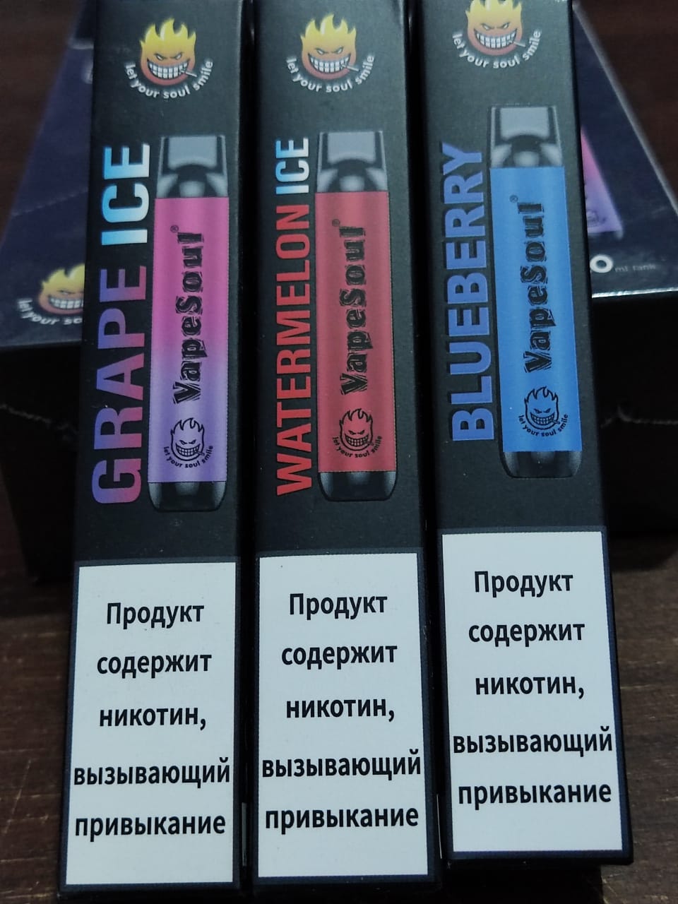 VapeSoul disposable vape devices in black, purple, and blue colors with flavor labels Grape Ice, Watermelon Ice, and Blueberry respectively, against a patterned background.